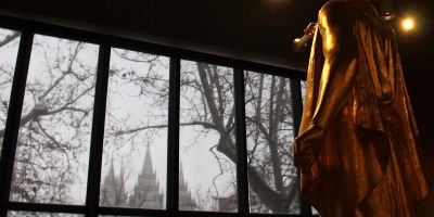 An Angel Moroni statue on display indoors looks out the window toward the Salt Lake Temple