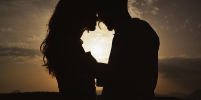 silhouette of a couple embracing, about to kiss, backed by the sun low in a partly cloudy sky